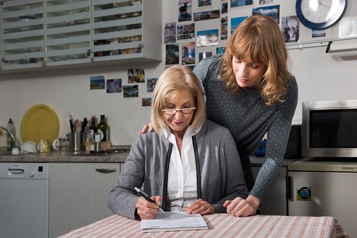 Eine bebrillte, ältere Frau Tisch sitzend füllt ein Formular aus, eine jüngere Frau hinter ihr stehend schaut ihr über die Schulter, eine Hand hat die stehende Frau der sitzenden Frau auf die rechte Schulter gelegt .