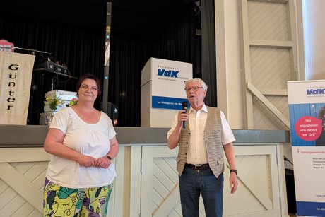 Eine Frau im T-Shirt und bunter Hose steht neben einem älteren Herrn im weißen Kurzarmhemd und Weste.
