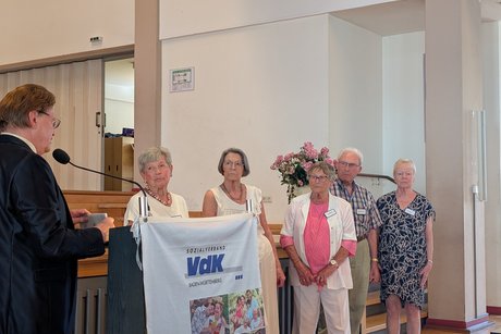 Mann am Rednerpult, seitlich vier ältere Frauen und ein älterer Herr, die ihm aufmerksam zuhören.