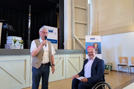 Links steht ein Mann im weißen Kurzarmhemd mit Mikrofon in der Hand. Daneben sitzt ein Rollstuhlfahrer im Anzug ohne Krawatte.