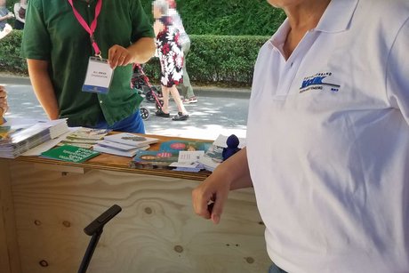 Eine Frau mit VdK-Poloshirt posiert vorne am Stand, im Hintergrund liest ein Mann im grünen T-Shirt die am Stand ausgelegten Broschüren.