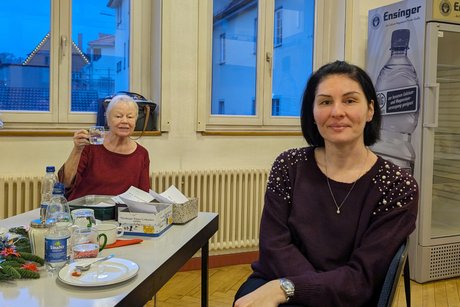 Zwei Damen an einem Tisch: Eine, näher an der Kamera, lächelt dezent in die Kamera, während die andere im Hintergrund ihr Glas in Richtung Kamera hebt.