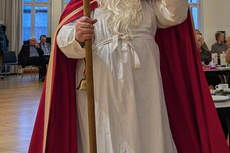 Vorweihnachtlicher Nachmittag Mann im Nikolauskostüm winkt mit einer Hand zur Kamera und hält in der anderen einen Bischofsstab.