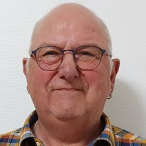 Etwas kr&auml;ftiger Mann im fortgeschrittenen Alter. Ein paar graue Haare schlie&szlig;en die sehr hohe Stirn ab. Er tr&auml;gt links einen silbernen Ohrring und eine dunkelgrau blaue Brille. Das ganze wird durch karrirtes Hemd abgerundet.