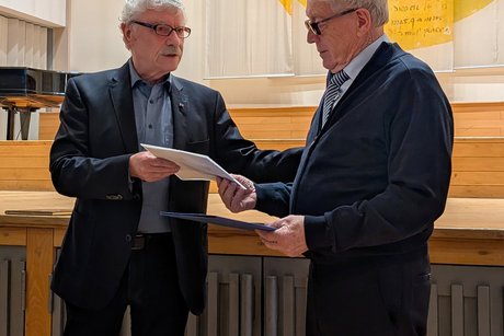 Vorweihnachtlicher Nachmittag Älterer Mann im Anzug überreicht einem weiteren älteren Mann im Anzug eine Urkunde.