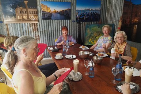Fünf Frauen sitzen zusammen und genießen Kaffee und Kuchen; an der Wand hängen Fotos von touristischen Sehenswürdigkeiten.