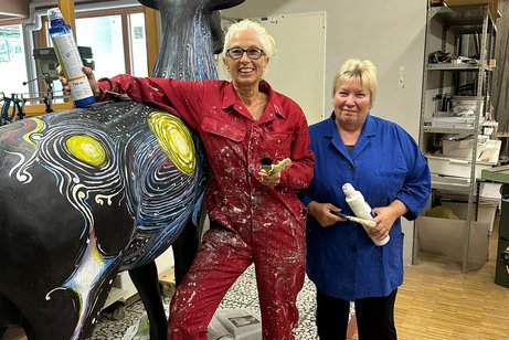 Angela Gutmann in rotem Blaumann und Ursula Karle mit dunkelblauem Oberteil, stehen malbereit neben der Hirschskulptur, die von hinten zu sehen ist.