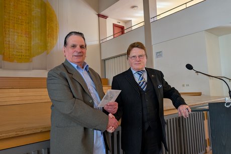 Zwei &auml;ltere, gut gekleidete Herren, die sich die H&auml;nde sch&uuml;tteln und in die Kamera blicken; einer der M&auml;nner h&auml;lt einen Umschlag in der Hand.