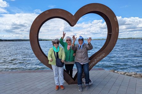 Drei &auml;ltere Damen stehen vor einer herzf&ouml;rmigen Metallskulptur am Ufer des Altm&uuml;hlsees, im Hintergrund der See.