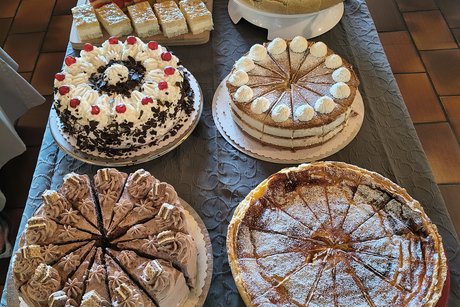 Mehrere verschiedene Kuchen und Torten auf einem dunklen Tisch, darunter runde Torten mit Sahne und Beeren sowie rechteckige Kuchenst&uuml;cke mit roten und gelben Bel&auml;gen.