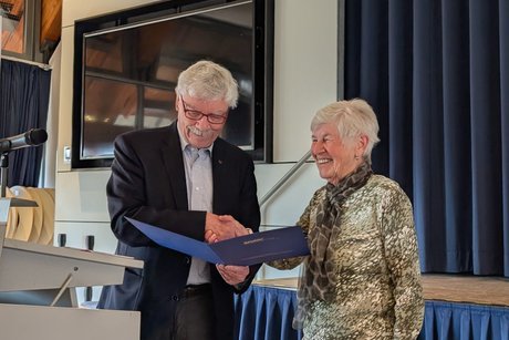 Ein elegant gekleideter Mann im Anzug und Hemd verliest eine Urkunde und sch&uuml;ttelt dabei die Hand einer elegant gekleideten &auml;lteren Dame.
