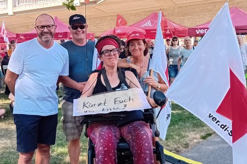 Freiburgs Sozialb&uuml;rgermeister Roland Meder, Schatzmeister Andreas Miller, VdK Dreisamtal, stehend von links Homepagebeauftragte Karin Maier im Elektrorollstuhl, mit umh&auml;ngendem Pappschild "K&uuml;rzt euch ins Knie" und ver:di-Fahne, Ortsvorsitzende Brigitte Mauz, stehend, im St&uuml;hlinger-Park in Freiburg