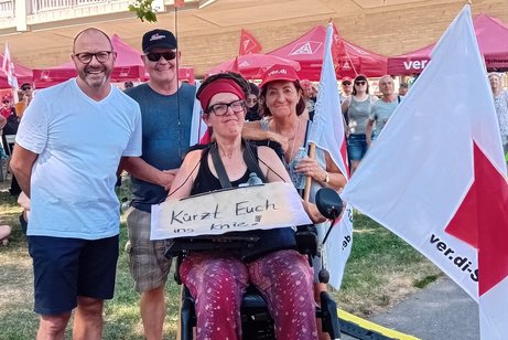 Freiburgs Sozialb&uuml;rgermeister Roland Meder, Schatzmeister Andreas Miller, VdK Dreisamtal, stehend von links Homepagebeauftragte Karin Maier im Elektrorollstuhl, mit umh&auml;ngendem Pappschild "K&uuml;rzt euch ins Knie" und ver:di-Fahne, Ortsvorsitzende Brigitte Mauz, stehend, im St&uuml;hlinger-Park in Freiburg