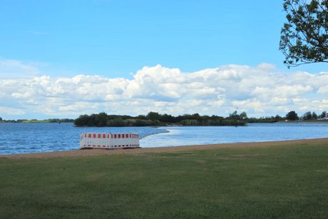 Panoramablick auf den durch einen schwimmenden Damm in zwei H&auml;lften geteilten See, mit gekr&auml;uselter Wasseroberfl&auml;che auf der linken und ruhiger, glatter Wasserfl&auml;che auf der rechten Seite.