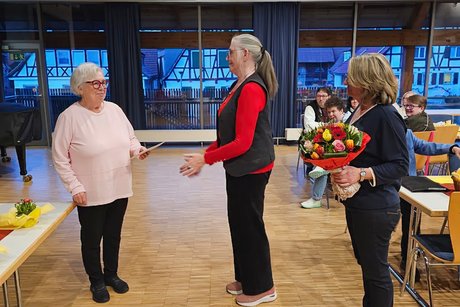 Hauptversammlung VdK OV Echterdingen 2026 Drei Damen: Die mittlere Dame geht auf die linke Dame zu, um sie zu umarmen, während die rechte Dame einen großen Blumenstrauß hält