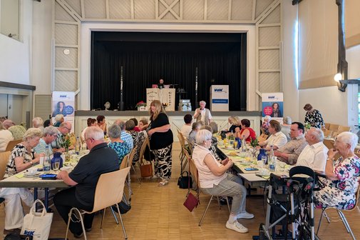 Großer Festsaal mit mehreren Tischreihen, an denen vorwiegend ältere Menschen sitzen. Im Hintergrund befindet sich eine Bühne, auf der ein DJ an seinem aufgebauten Equipment sitzt.