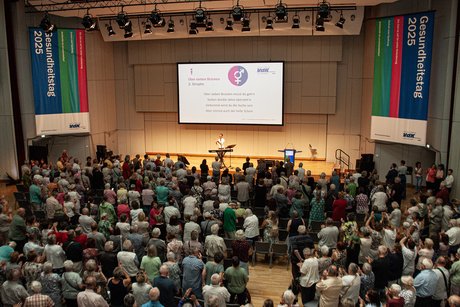 Ein Teil der Zuhörer, alle stehen, singen und schauen auf die Leinwand vorne an der Bühne, dort ist der Text zu dem Lied "Über sieben Brücken" zusehen, links und rechts von der Bühne stehen VdK-Fahnen