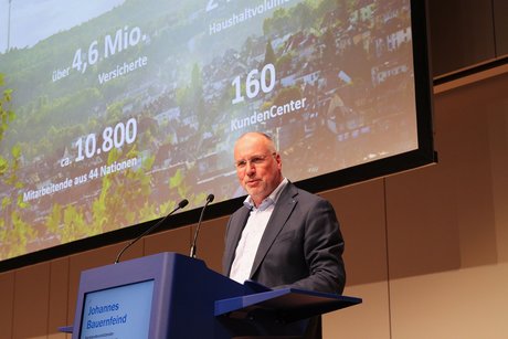 Johannes Bauernfeind, Vorstandsvorsitzender der AOK Baden-Württemberg steht am Pult und am Mikrofon und spricht, hinter ihm diverse Zahlen an der Leinwand