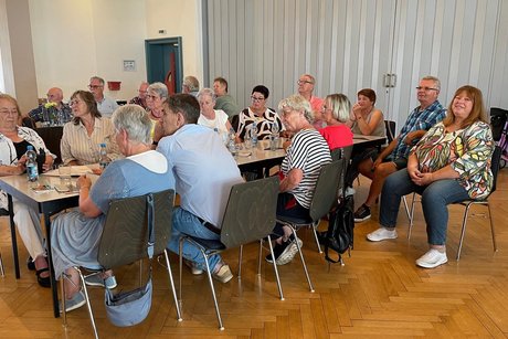 Ein voll besetzter Tisch mit einer bunt gemischten Runde von Menschen, die dicht beieinandersitzen und aufmerksam den Blick zur B&uuml;hne richten.