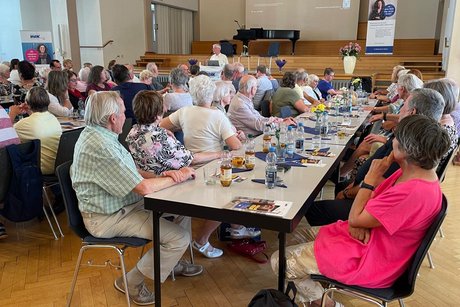 Panorama des Festsaals mit drei Tischreihen, an denen ein bunt gemischtes Publikum sitzt und aufmerksam einem Vortrag lauscht.