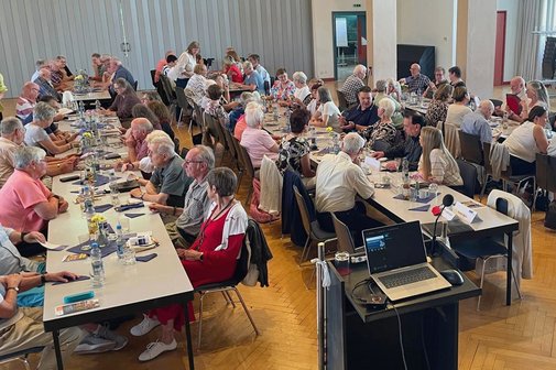 Panorama des Festsaals mit drei Tischreihen, an denen eine bunt gemischte, fr&ouml;hliche Runde von Menschen sitzt und sich angeregt unterh&auml;lt. Zentral im Bild ein Rednerpult mit ge&ouml;ffnetem Laptop.