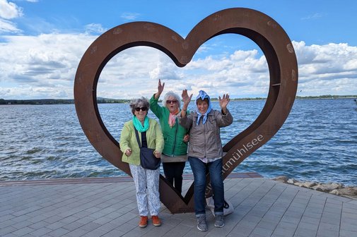Drei &auml;ltere Damen stehen vor einer herzf&ouml;rmigen Metallskulptur am Ufer des Altm&uuml;hlsees, im Hintergrund der See.