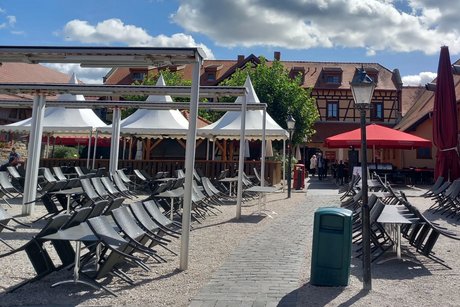 Au&szlig;enbereich mit Kiesfl&auml;che, mehreren Tischen und St&uuml;hlen, teilweise unter Sonnenschirmen; die Tische sind unbesetzt, die St&uuml;hle an die Tische gelehnt.