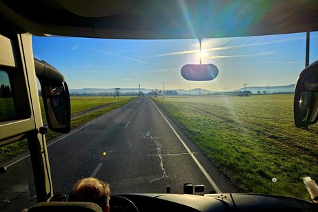 Durch die riesige Windschutzscheibe des Reisebusses sieht man die Sonne &uuml;ber taugr&uuml;nen Wiesen.