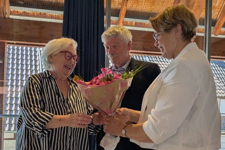 Eine Dame &uuml;berreicht einer anderen Dame einen Blumenstrau&szlig;, die l&auml;chelnd die &Uuml;bergabe entgegennimmt. Daneben steht ein Mann, der die Szene beobachtet.