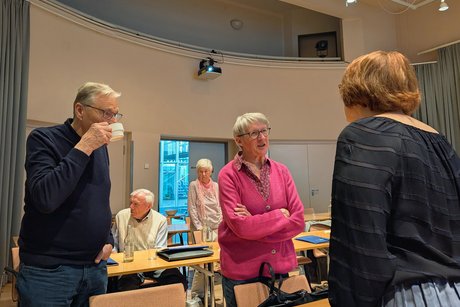 Zwei Frauen unterhalten sich stehend; daneben steht ein Mann, der zuhört und eine Tasse Kaffee trinkt.