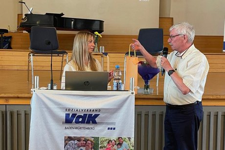 Dame steht hinter einem Rednerpult. Ein &auml;lterer Mann neben ihr &uuml;berreicht ihr eine Geschenktasche mit Sekt.