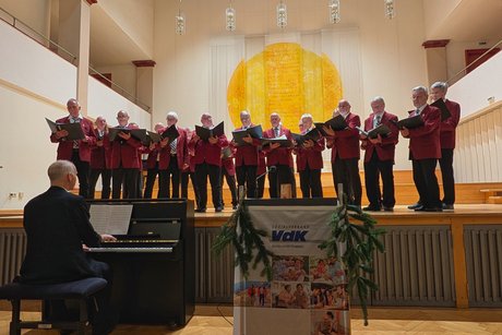 Vorweihnachtlicher Nachmittag Rund 15 Männer in schwarzen Hosen und roten Sakkos singen auf der Bühne, davor sitzt ein Mann im schwarzen Sakko am Klavier und spielt.