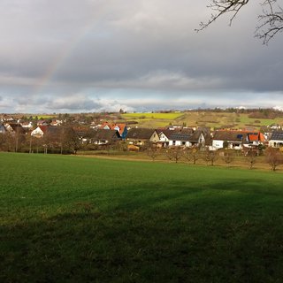 Mittig im Tal liegend Häuser von Gräfenhausen. Im Vordergrund ein weiter grüner Acker, teils sonnenbeschienen, teils schattig. Am Himmel dichte Wolken und ein Regenbogen.