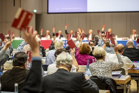Innenaufnahme der Liederhalle in Stuttgart, Veranstaltungssaal, Nhaufnahme von hinten auf die Delegierten des Sozialverbandes VdK, ausgestreckte H&auml;nde sind zu sehen, die rote Stimmk&auml;rtchen halten, die Delegierten reihen sich auf ihren Pl&auml;tzen aneinander, der Saal ist voll. 