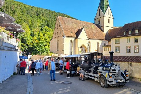 Touristenbahn als Mini-Dampfloko vor einer Kirche mit Turm in einer Dorfstra&szlig;e. Menschen warten am Zug, bewaldeter H&uuml;gel im Hintergrund.