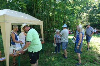 Grillstation und Essensausgabe im Garten des Winzermuseums. Das Vorstandsmitglied Hannelore M. steht unter dem Pavillion beim Grill vor einen Biertisch für die Essensausgabe. Mehrere Gäste stehen an und warten auf ihre Bestellung. Im Hintergrund ist ein Schuppen zu sehen