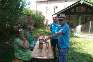 Die Kassierin Silvia S. und Vorstandsmitglied Karin B. sitzen im Garten des Winzermuseums auf einer Bierbank an der Bon-Kasse, die auf dem Biertisch vor ihnen steht. Sie sind im Gespräch mit Peter F. und einem Gast. Im Hintergrund ist ein grüner Schuppen zu sehen