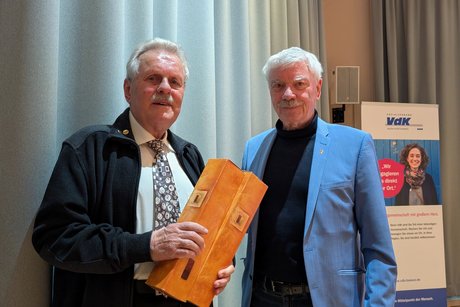 Zwei ältere Männer stehen nebeneinander und lächeln in die Kamera; der Mann links hält zwei Geschenkkartons mit Wein.