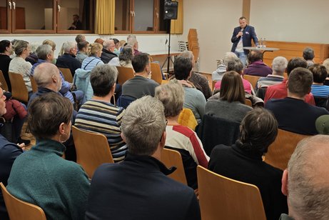 Besuchende auf St&uuml;hlen in Stuhlreihen sitzend beim Vortrag von Dr. Fabian Hetmeier (vorne) in Pfarrsaal