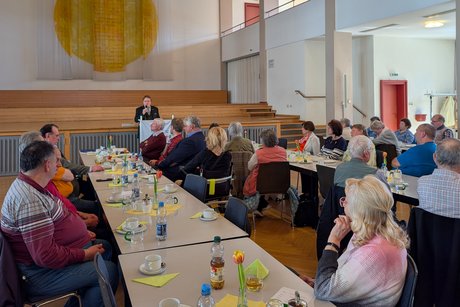 Im Vordergrund zwei Tischreihen mit sitzenden Menschen, die aufmerksam dem Redner am Pult vorne lauschen.