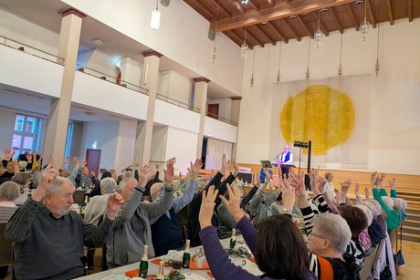 Tischreihen im Festsaal, während die Gäste im Rhythmus der Musik beide Arme in die Luft strecken.