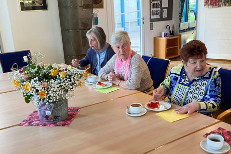 Drei Damen sitzen an einem Tisch bei Kaffee und Erdbeerkuchen und unterhalten sich; in der Mitte steht ein prächtiger Blumenstrauß.