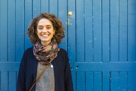 Junge Frau mit lockigen Haaren und breitem Lachen strahlt in die Kamera, sie steht vor einem Holzzaun, der blau gestrichen ist.
