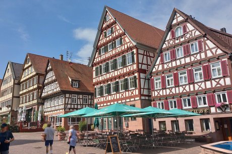 Marktplatz mit vier Fachwerkhäusern und teilweise davor stehenden Tischen mit Sonnenschirmen der Außengastronomie.