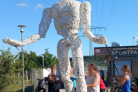 Drei Personen führen eine etwa 3 Meter große weiße Marionettenfigur an langen Stäben.