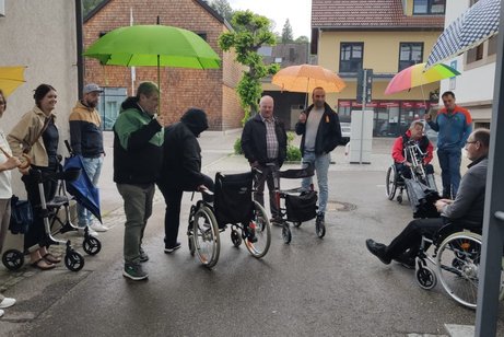 Für viele eine neue Erfahrung, ein noch zu besetzender Rollstuhl, einige schon besetzte Rollstühle, eine Person am Rollator, mehrere Menschen mit Schirmen vor dem neuen MTB-Haus