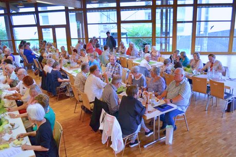 Panorama des Festsaals mit mehreren Tischen, an denen Menschen sitzen und vespern.