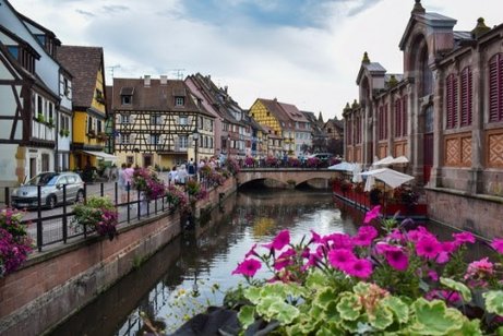 Blick von einer Br&uuml;cke auf einen malerischen Ort im Elsass mit alten Fachwerkh&auml;usern, Bachlauf und bunt bl&uuml;henden Blumen im Vordergrund.