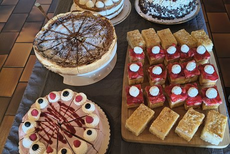 Mehrere verschiedene Kuchen und Torten auf einem dunklen Tisch, darunter runde Torten mit Sahne und Beeren sowie rechteckige Kuchenst&uuml;cke mit roten und gelben Bel&auml;gen.