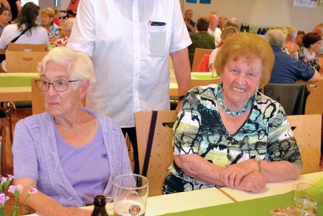 Zwei Frauen am Tisch sitzend, dahinter ein stehender Mann. Alle lächeln in die Kamera.